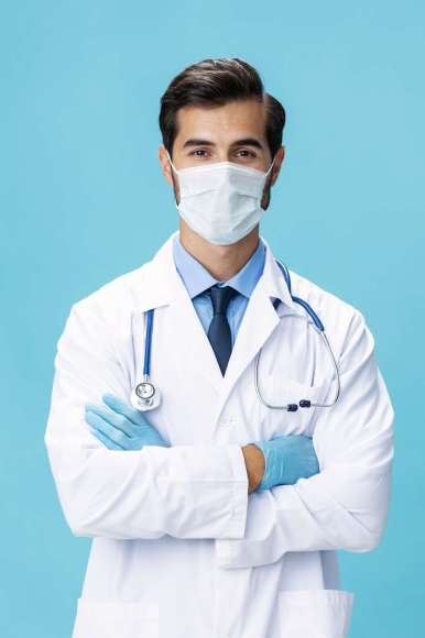 A male doctor in a white coat and a medical mask stands with his hands and looks at the camera on a blue isolated background, copy space, space for text. High quality photo